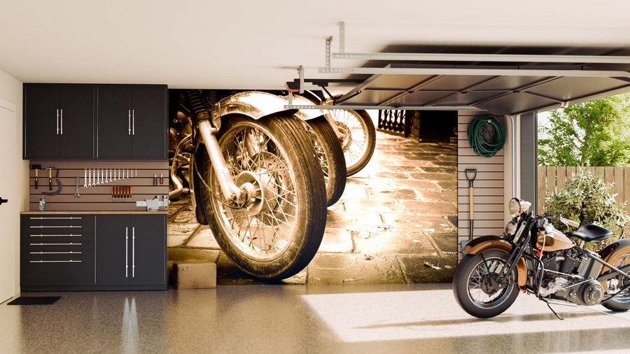 A clean, modern garage with black cabinets and organized tools features the Bikes in a Row Wall Mural of vintage motorcycle wheels on the back wall. A classic motorcycle is parked inside as sunlight streams in, revealing greenery outside.