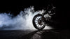 The Motorcycle Burnout Smoke Wall Mural from Deposit Photo depicts a dramatic scene of a motorcycle performing a burnout on a dark surface, emitting thick white smoke. The prominent front wheel and reflective rim create striking contrast against the black background.