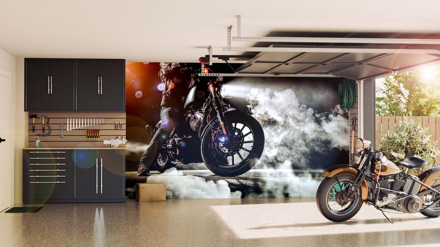 A tidy garage with black storage cabinets and tools on the left. A motorcycle is parked on the right, while the back wall features the Foggy Motorcycle Wall Mural, depicting a motorcycle in action amid swirling smoke. Sunlight enters from an open door.