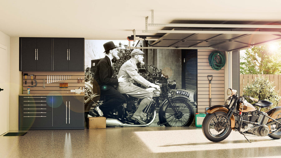 A modern, tidy garage features black cabinets, organized tools, and a classic motorcycle. The focal point is the Men Riding Motorbike Wall Mural—a large black-and-white image of two men on a vintage motorcycle. Sunlight streams in from the open garage door.