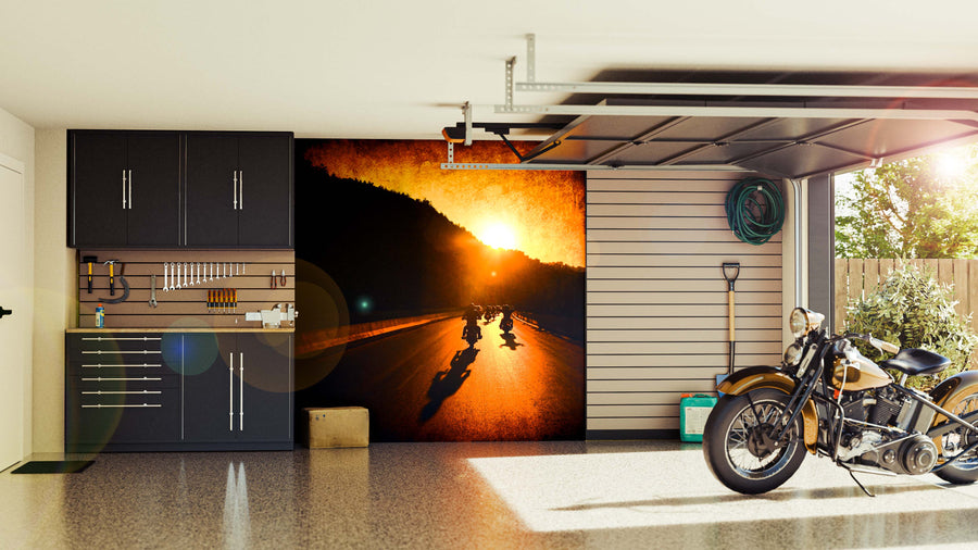 A sleek garage features black cabinets, tidy tools, a vintage motorcycle, and the “Motorcycle Ride at Sunset Wall Mural,” depicting bikers cruising at sunset. Sunlight streams through the open door onto the polished floor.
