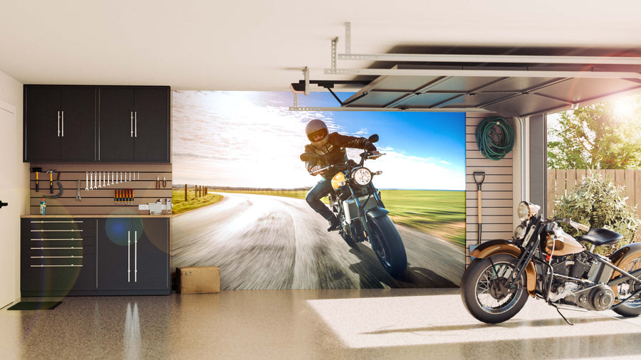 A tidy garage features gray cabinets, tools arranged on wall racks, a motorcycle on a speckled floor, and a large Motorcycle Rider Adventure Wall Mural showing someone riding down a sunny rural road as sunlight pours in through the open door.