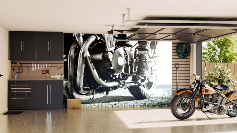 A clean, modern garage features dark cabinets, organized tools, a coiled green hose, and a large Motorcycle Rider Boot Wall Mural. A classic brown and black motorcycle rests on the shiny, speckled floor near the open garage door.