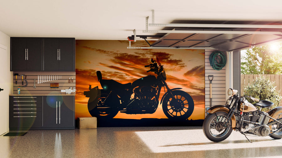 A modern garage showcases sleek black cabinets, a motorcycle on a shiny floor, and the Motorcycle Silhouette Wall Mural—a vibrant display of a bike against an orange sunset—while sunlight streams in from the open door.
