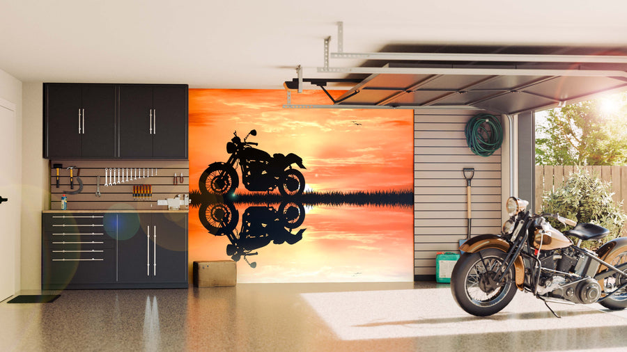 A modern garage with black cabinets, organized tools, and a classic motorcycle features the Motorcycle at Sunset Wall Mural—a peel and stick mural depicting a motorcycle’s silhouette on water framed by a vivid orange sunset.