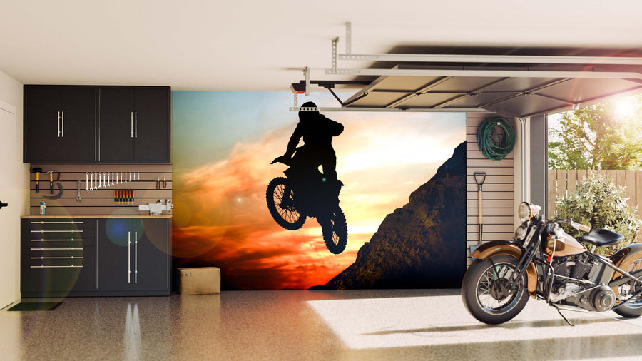 A modern garage with dark cabinets and a motorcycle parked inside features the "Motorcyclist Jump Off Cliff Wall Mural," showcasing a silhouette of a rider leaping against an orange sunset, lit by sunlight through the partly open garage door.