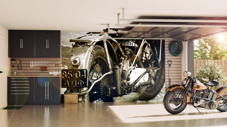 A modern garage with sleek black cabinets and organized tools features the Old Motorcycle Engine Wall Mural in sepia tones on the back wall, highlighted by sunlight streaming through the open door and a vintage motorcycle parked inside.