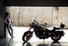 A woman in plaid and ripped jeans leans against a garage door, admiring a red motorcycle inside. The sunlight highlights the bike's sleek design, evoking the "Motorcycle and Rider Wall Mural" by Deposit Photo, known for its artful details in living spaces.