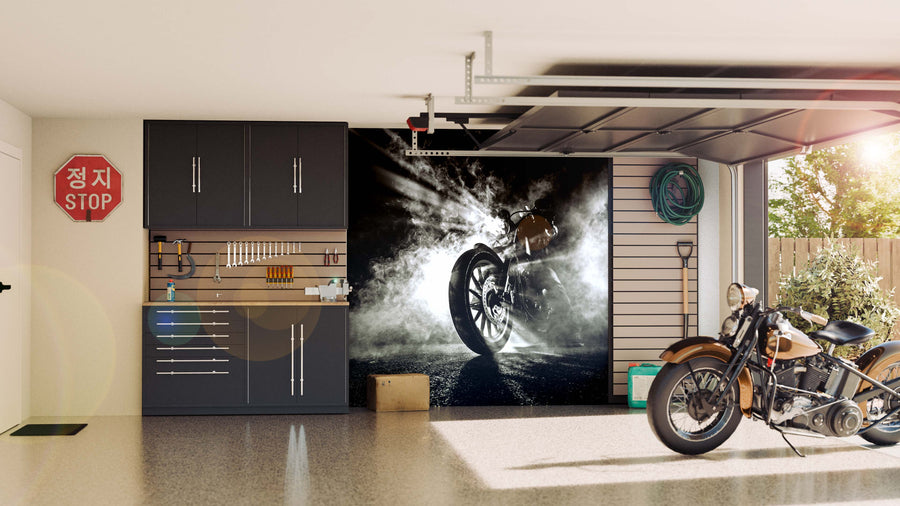 A tidy garage with black cabinets, tools on a wall rack, a hose, and a large Smoke Wall Mural. A vintage motorcycle sits on the shiny floor, sunlight streams in, and a Korean stop sign decorates the wall by the open door to the green yard.