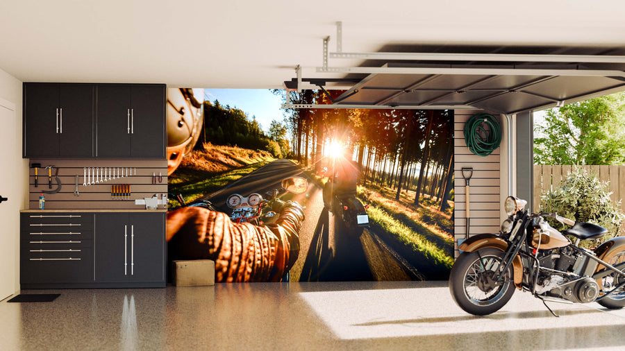 A clean, modern garage with dark cabinets, organized tools, and a classic motorcycle on a shiny floor features the Sunny Morning Ride Wall Mural—a scenic peel and stick mural showing sunlight streaming through trees on a country road.