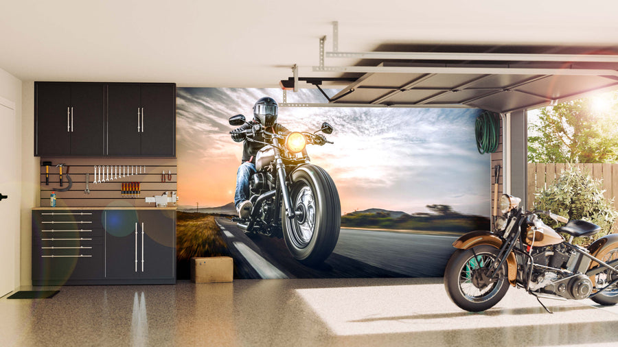 A tidy garage with dark cabinets and tools on the left, a classic motorcycle inside, and the Sunset Biker Wall Mural featuring a motorcyclist at sunset on the back wall. Natural light streams in through the open garage door.