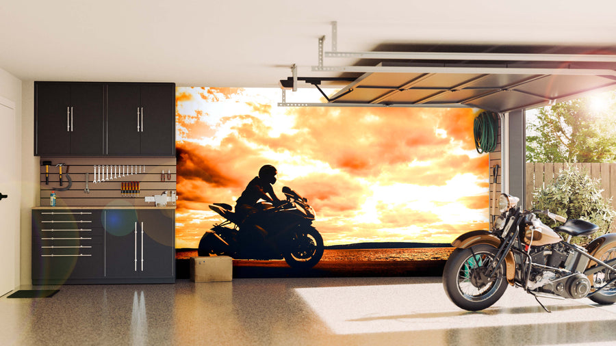 A modern garage with black cabinets, organized tools, and two motorcycles features the Sunset Chase Wall Mural—a striking image of a rider against a sunset sky—revealed through the open door as sunlight brightens the polished space.