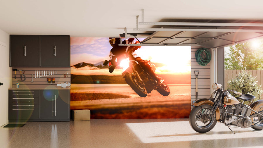 A modern garage features sleek cabinets, organized tools, and a classic motorcycle. The back wall displays the Sunset Drift Wall Mural, depicting a motorcyclist at sunset with warm sunlight creating a vibrant, dynamic atmosphere.