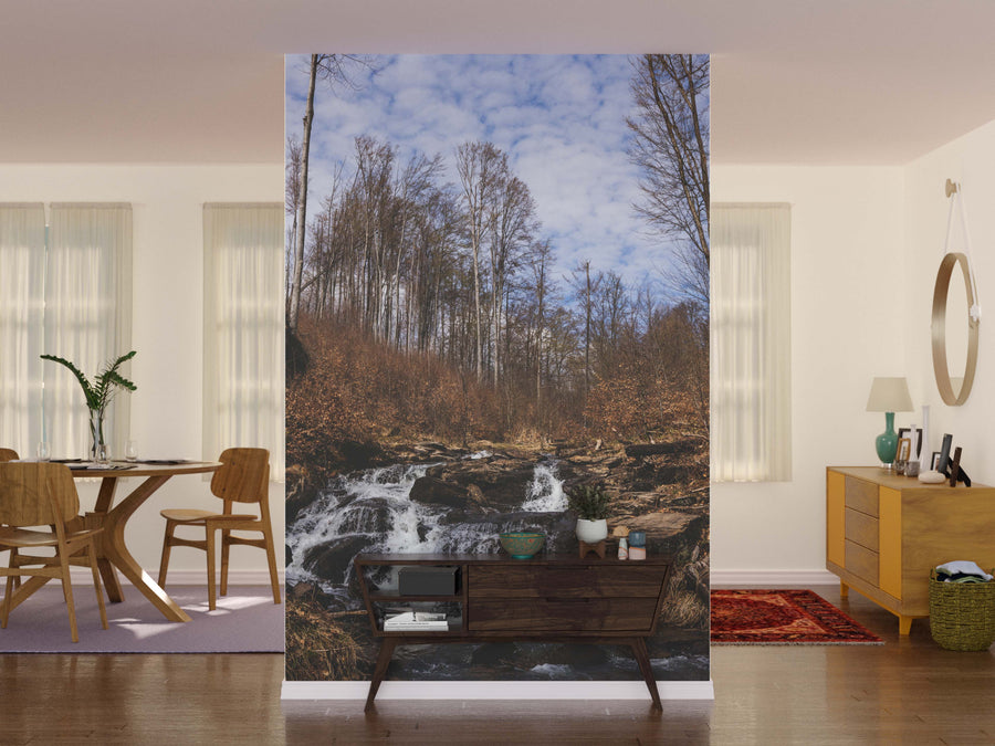 A dining area with a round table, chairs, and a wooden sideboard sits before the Mountain River with Stones Wall Mural, which depicts a forest stream beneath a cloudy sky and brings the beauty of nature indoors.
