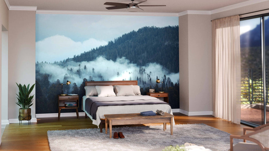 Modern living-dining room with wood floors, teal chairs, patterned rugs, and a large Mountain with Fog Wall Mural. Pendant lights hang above a dark dining table; the kitchen is visible behind a glass wall in this earthy space.
