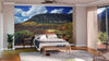 A modern bedroom features the Mountains Over Last Dollar Road Wall Mural—a large peel and stick mountain landscape with fall forest colors behind the bed—plus a bed, nightstands, bench, potted plant, rug, wood floor, ceiling fan, and balcony access.
