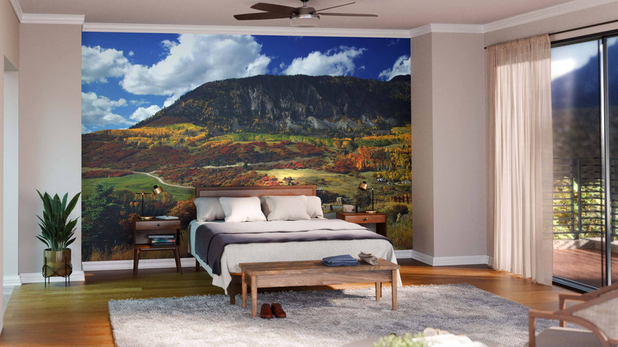 A modern bedroom features the Mountains Over Last Dollar Road Wall Mural—a large peel and stick mountain landscape with fall forest colors behind the bed—plus a bed, nightstands, bench, potted plant, rug, wood floor, ceiling fan, and balcony access.