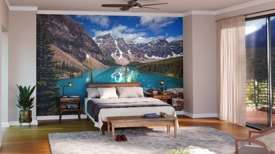 A modern bedroom features a large bed, potted plants, and a wooden bench. Sunlight warms the space through glass doors, highlighting the Mt. Allen and Moraine Lake Wall Mural of turquoise water and mountains behind the bed.