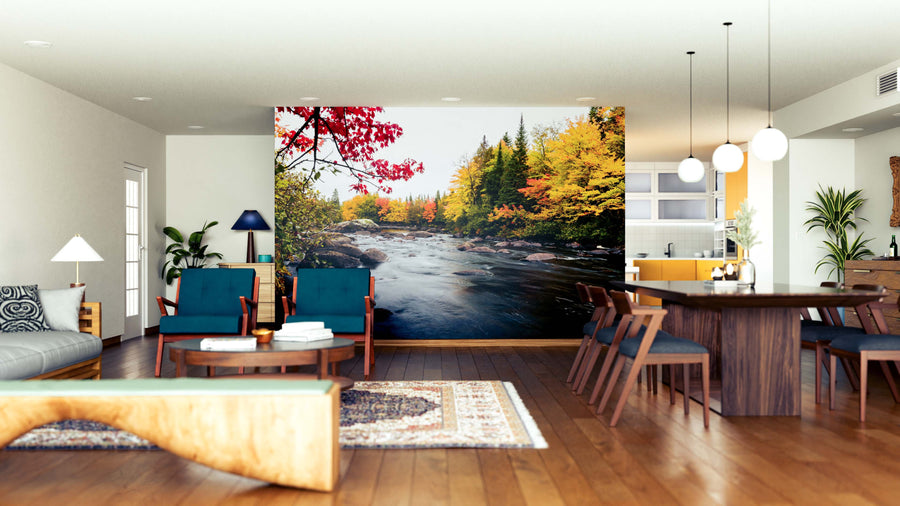 A modern open-plan living and dining room with wood floors, blue armchairs, and a wooden table features the North Quebec Wall Mural, showcasing a river and autumn forest in vibrant red, yellow, and green tones.