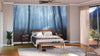 A cozy bedroom features a large bed with white and gray bedding, wood furniture, a potted plant, a gray rug, and the November Morning Misty Forest Wall Mural behind the bed, softly lit by sunlight through sheer-curtained glass doors.