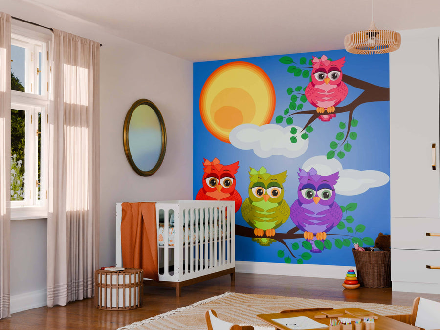 A nursery featuring a crib, wicker hamper, toys, and the Four Vibrant Owls Wall Mural showing cartoon owls on branches against a blue sky. Light pours in from a window; a round mirror and pendant light finish the look.