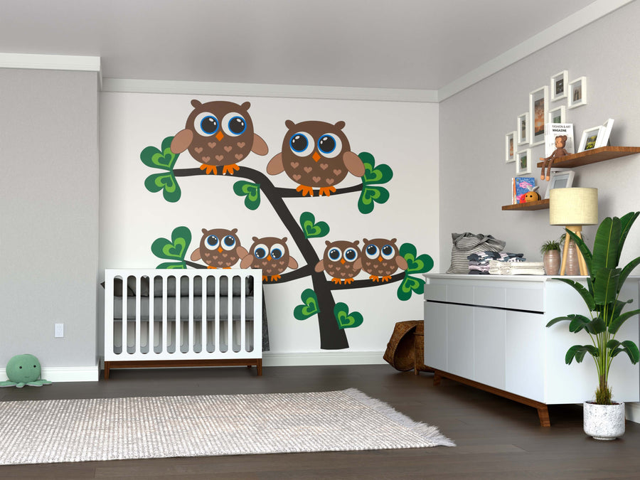 A modern nursery includes a white crib and dresser, brightened by the “An Owl Family Wall Mural” featuring five cartoon owls on a leafy tree. Decor and toys fill shelves, with a green plush toy on the hardwood floor for a cozy touch.