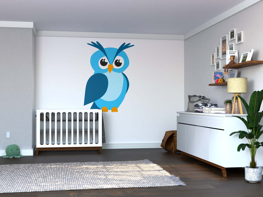 A modern nursery includes a crib, dresser with baby supplies, wooden floor, gray rug, green plant, framed pictures on a shelf, and playful decor featuring the Blue Cartoon Owl Wall Mural. The simple design uses neutral tones with fun accents.