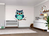 A modern nursery features a crib, white dresser, potted plant, and wall shelves. A large Blue Owl Wall Mural decorates the space above a branch. A light rug covers the wooden floor, with framed photos and stuffed toys on the shelves and dresser.