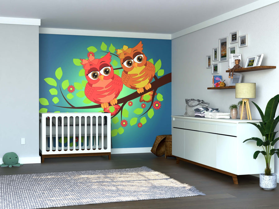 A nursery with a crib, dresser, and the Bright Owls Wall Mural showing two colorful cartoon owls on a tree branch. The room includes decorative frames, a plant, basket, soft toy, wood floors, and neutral-toned walls and ceiling.