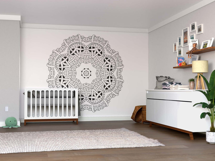 Modern nursery with a white crib, soft rug over wood floors, white dresser, shelf with toys and photos, green plant, basket, and a striking Color My Binded Mandala Wall Mural - Peel and Stick Wall as a custom black-and-white mural centerpiece.