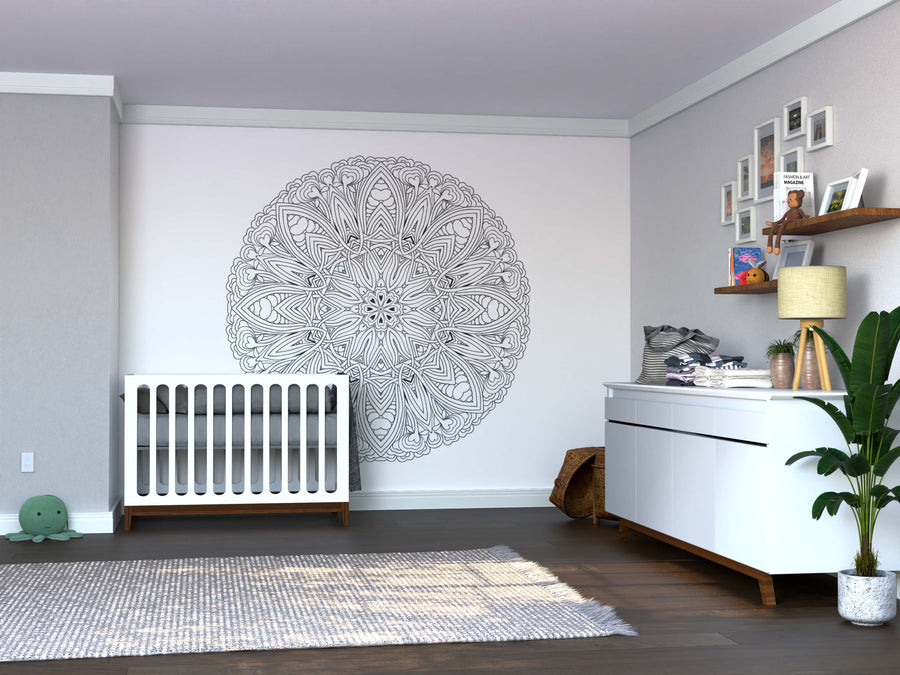 Modern nursery with gray walls, white crib and dresser. The wall behind the crib features the Color My Celtic Mandala Wall Mural. There’s a shelf with pictures and toys, potted plant, gray rug, and a basket with blankets on the wooden floor.