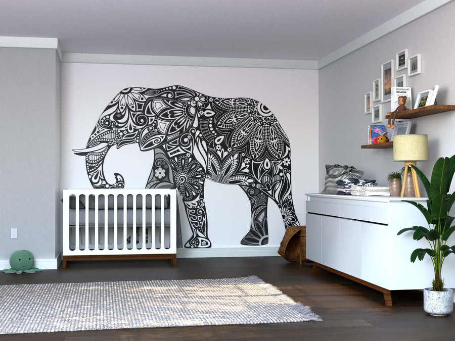 A modern nursery with a white crib, dresser, and shelves features the Color My Elephant Wall Mural in black and white. A potted plant, framed photos, baby supplies, and a plush turtle add warmth alongside wooden and gray accents.