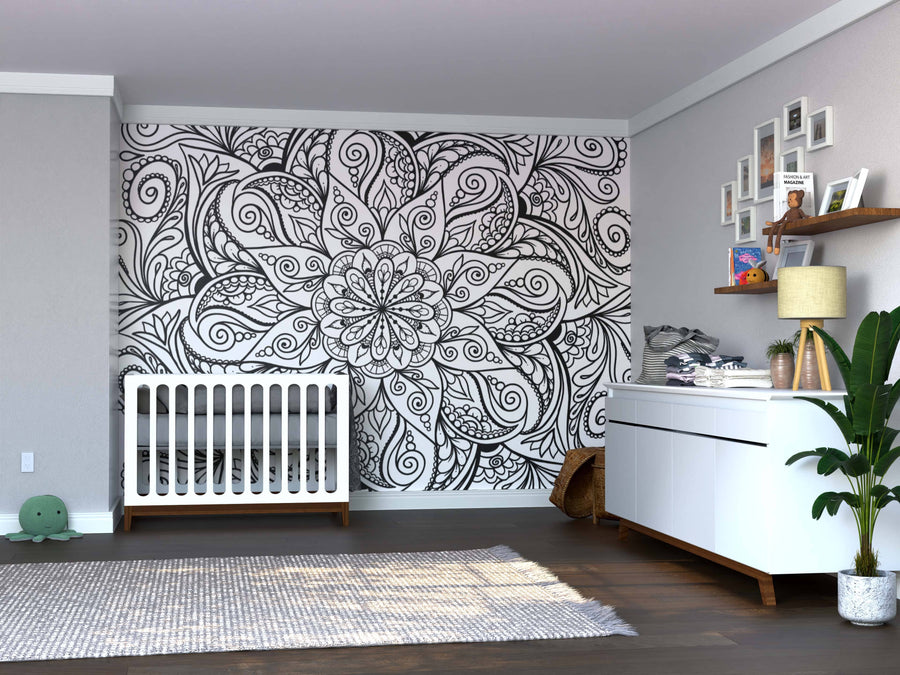 A nursery with a white crib, dresser with lamp and baskets, and the Color My Floral Maze Wall Mural as a striking black-and-white peel and stick backdrop. Gray walls, wood floors, photo shelves, a green plant, and gray rug add cozy finishing touches.