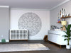 A modern nursery features a gray crib, dresser with baby supplies, lamp, and shelves. Above the crib, the Color My Spade Mandala Wall Mural adds style. A rug, leafy plant, and framed photos decorate the shelves.