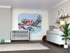 A nursery with a white crib, dresser, plant, baby items, and the A Colorful Owl Family Wall Mural featuring cartoon owls on tree branches. The room has soft gray walls, wooden floors, and a small green turtle toy on the rug.