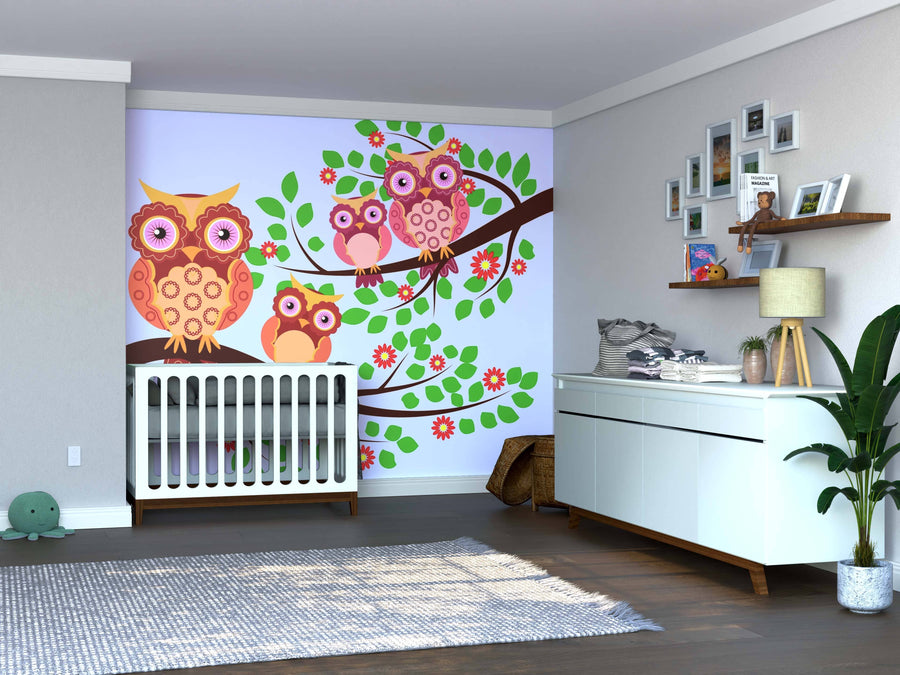 A modern nursery with light walls and wooden floors centers on a crib before the Colorful Owls on Flowered Tree Wall Mural. A white dresser, potted plant, and gallery wall of framed photos complete the inviting space.