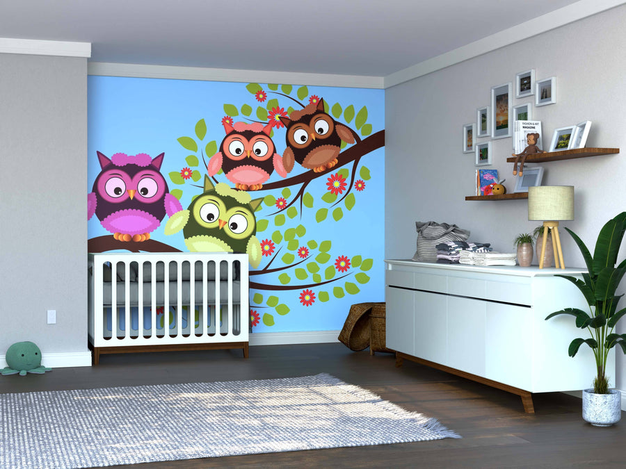 A nursery with a white crib and the Colorful Owl Friends Wall Mural featuring four cartoon owls on a tree, plus a white dresser with decor, framed photos, a green plant, wooden floors, and a plush turtle toy on the floor.