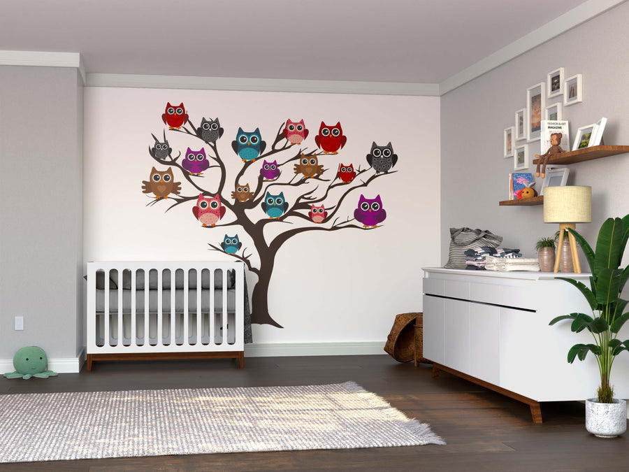 A nursery features a white crib, dresser, potted plant, and the Colorful Owl Tree Wall Mural. Dark wood floors, a green plush toy in the corner, and framed photos on shelves add charm to this inviting space.