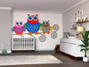 A nursery room with a crib, dresser, and houseplant. Above the crib, the Colorful Owls Wall Mural—a peel and stick design featuring three cartoon owls on a branch—adds charm. Framed pictures and plush toys complete the playful decor.