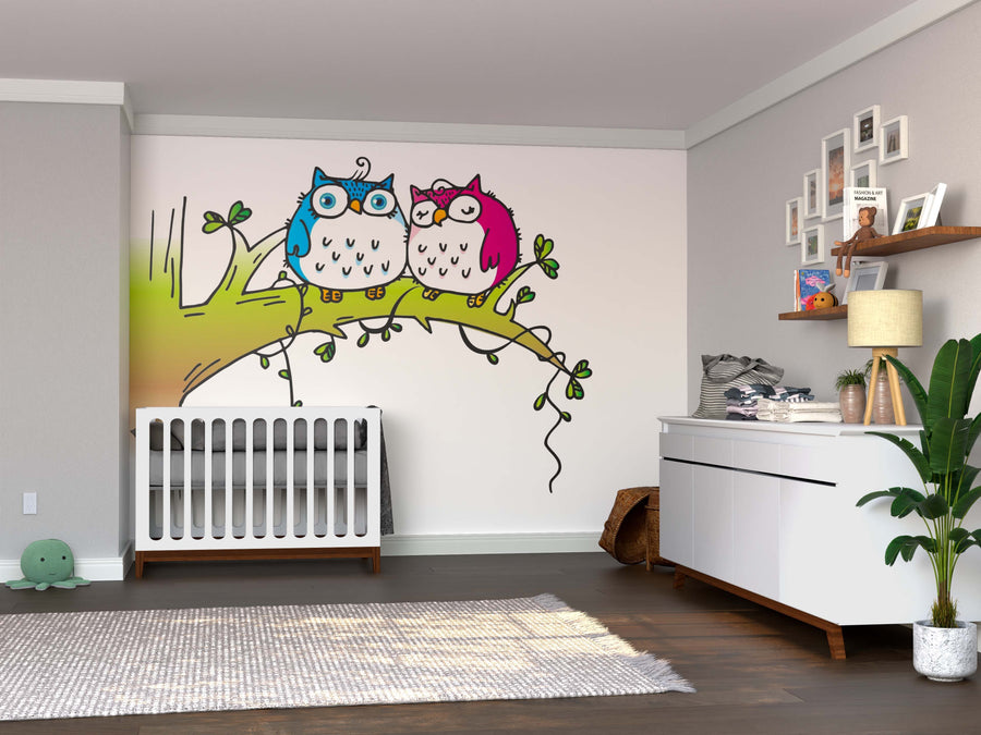 A nursery features a crib, dresser, plant, and the Couple Owl Wall Mural with two cartoon owls—blue and pink—on a tree branch. Removable wallpaper adds charm, while shelves above the dark wood floor display books and decor beside a green plush toy.