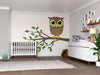 A modern nursery has a crib on the left, a Cute Baby Owl Wall Mural on a tree branch, a white dresser with toys and plants, framed photos above, and a wooden floor with a green plush turtle toy in the corner.