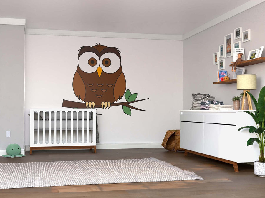 A nursery room with a white crib, dresser, shelves with decor, and the Cute Brown Owl Wall Mural depicting a cartoon owl on a branch. The space features light walls, wooden floors, a gray rug, green plant, and framed photos above the dresser.