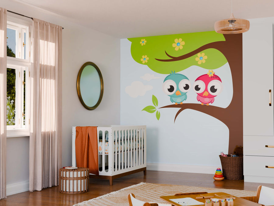 A nursery with a crib, ottoman, rug, toys, and mirror. The wall displays the Cute Love Birds Wall Mural featuring two cartoon owls—one blue, one pink—on a leafy branch with yellow flowers as sunlight streams in.