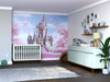 A nursery with a white crib, dresser, and shelves features the Enchanted Pink Castle Wall Mural with butterflies and trees. A gray rug covers the wooden floor, accented by toys, plants, and framed pictures on walls and shelves.