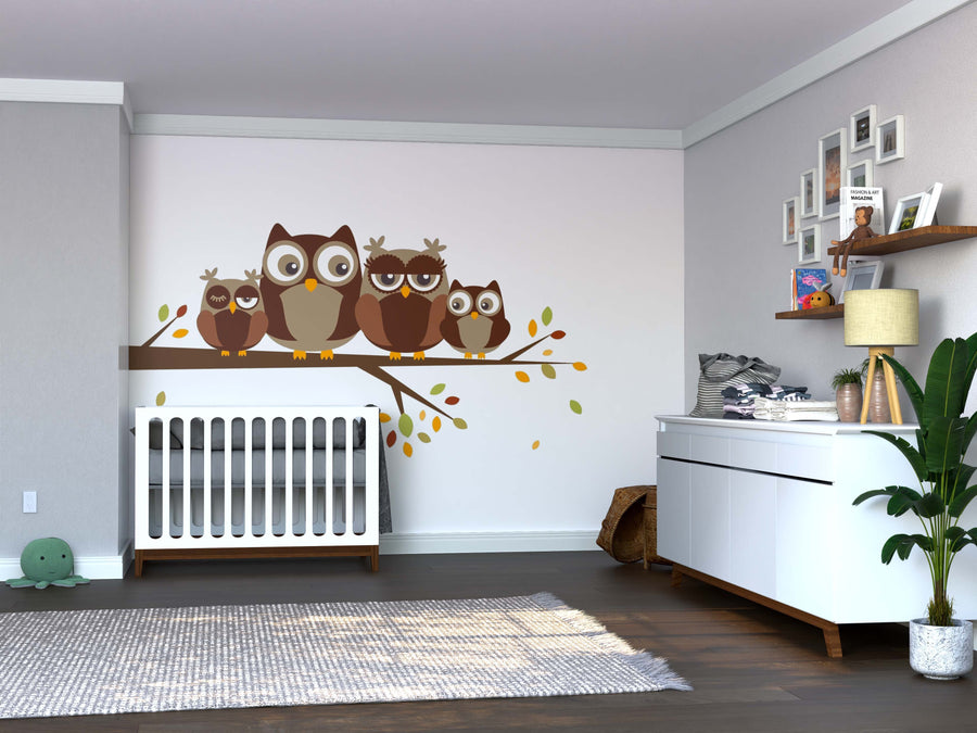 A modern nursery features a white crib, dresser, and plant. Above the crib, the Family of Owls Wall Mural with four brown owls on a branch adds charm. Shelves and framed pictures complete the cozy decor.
