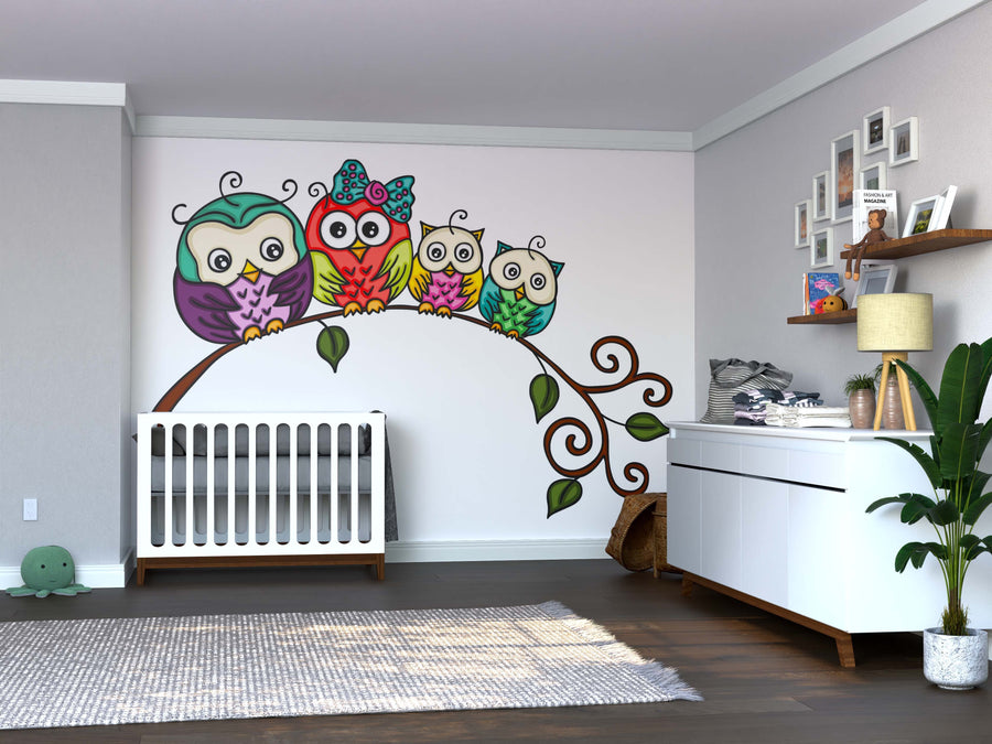 A modern nursery with a crib, white dresser, plant, and wooden floors features the Family of Colorful Owls Wall Mural—four cartoon owls on a branch. Above the dresser, shelves display baby items and framed photos.
