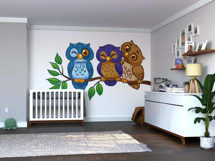 A nursery with a white crib, dresser, gray rug, toys, framed art, plants, and a shelf with baby items feels cozy. The highlight is the Friendly Owls Wall Mural—three cartoon owls on a branch in peel-and-stick style.