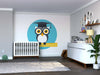 A modern nursery with light gray walls, a white crib, and dresser with changing pad. The Graduating Owl Wall Mural shows an owl in a graduation cap on a ruler. Shelves display books and framed pictures above the dresser.
