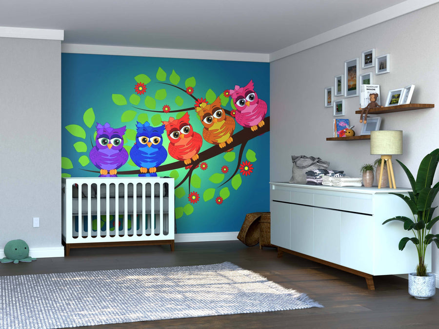 A nursery features a white crib against a blue wall decorated with the Group of Colorful Owls Wall Mural, showing five cartoon owls on a branch. The room also includes a white dresser, houseplants, wooden floor, shelves with decor, and streaming sunlight.