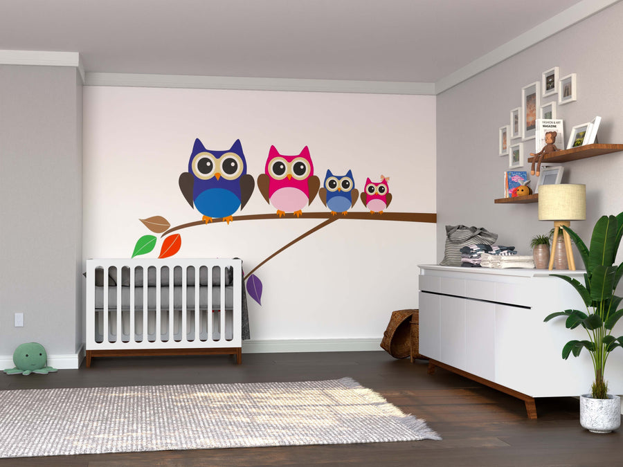 A nursery with a white crib, dresser, green plant, and neutral tones features the Happy Owl Family Wall Mural—four colorful owls on a branch—plus a cozy rug and framed photos above the dresser.